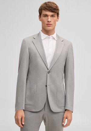 Mann trägt einen hellgrauen Zweiknopfanzug mit aufgesetzten Taschen über einem weißen Poloshirt mit Kragen und steht vor einem schlichten Hintergrund.