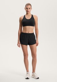Čierny športový podprsenka s okrúhlym výstrihom v kombinácii so voľnými čiernymi šortkami. Model nosí svetlé športové topánky. Hladká textúra látky.