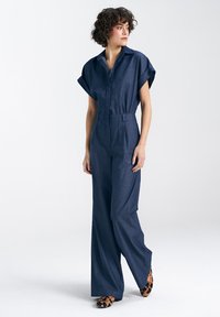 Jeansowy kombinezon z krótkimi rękawami, frontowymi guzikami, dopasowanym krojem, szerokimi nogawkami i paskiem w talii. Noszony z balerinami w panterkę.