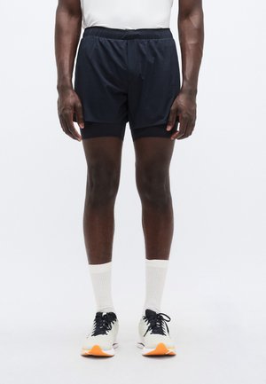 Vyriški tamsiai mėlyni sportiniai šortai su tampriu liemeniu, turintys integruotą juodą pamušalą ir lygią medžiagą. Derinami su baltais kulkšnies ilgio kojinėmis ir bėgimo bateliais.