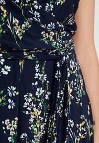 Marinblå blommig omlottklänning med vita och gröna blommönster. Mjuk, lätt vikt med knytband i midjan för justerbar passform.