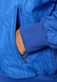 Plavi dukserica sa patentnim zatvaračem koja ima teksturirani print s logotipima. Tkanina je glatka, s rebrastim manžetama i džepom sa strane.