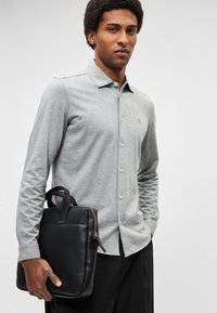 Camisa cinzenta de mangas compridas com botões, gola pontiaguda e um logótipo subtil, combinada com uma bolsa texturada de couro preto com alças duplas.