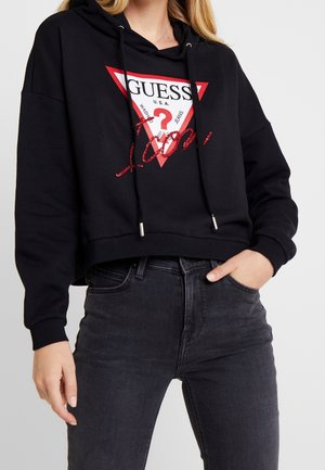 Femme portant un sweat à capuche court noir avec le logo Guess rouge et blanc et un jean gris foncé, main dans la poche, debout sur fond blanc.
