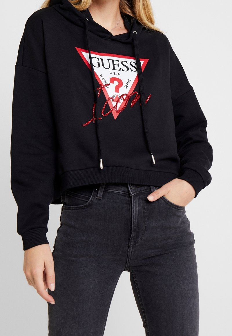 Femme portant un sweat à capuche court noir avec le logo Guess rouge et blanc et un jean gris foncé, main dans la poche, debout sur fond blanc.