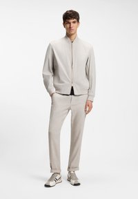 Joven de pie, vestido con una chaqueta beige clara con cremallera, pantalones a juego con puños y zapatillas beige con detalles en negro y blanco.