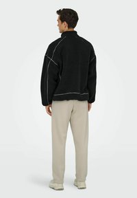 Black fleece jacket with contrasting white stitching, high collar, and relaxed fit, paired with light beige pants and neutral sneakers.