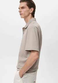 Beige kortärmad pikétröja med knäppbar krage, slät textur och figursydd passform, bärs med ljusa byxor.