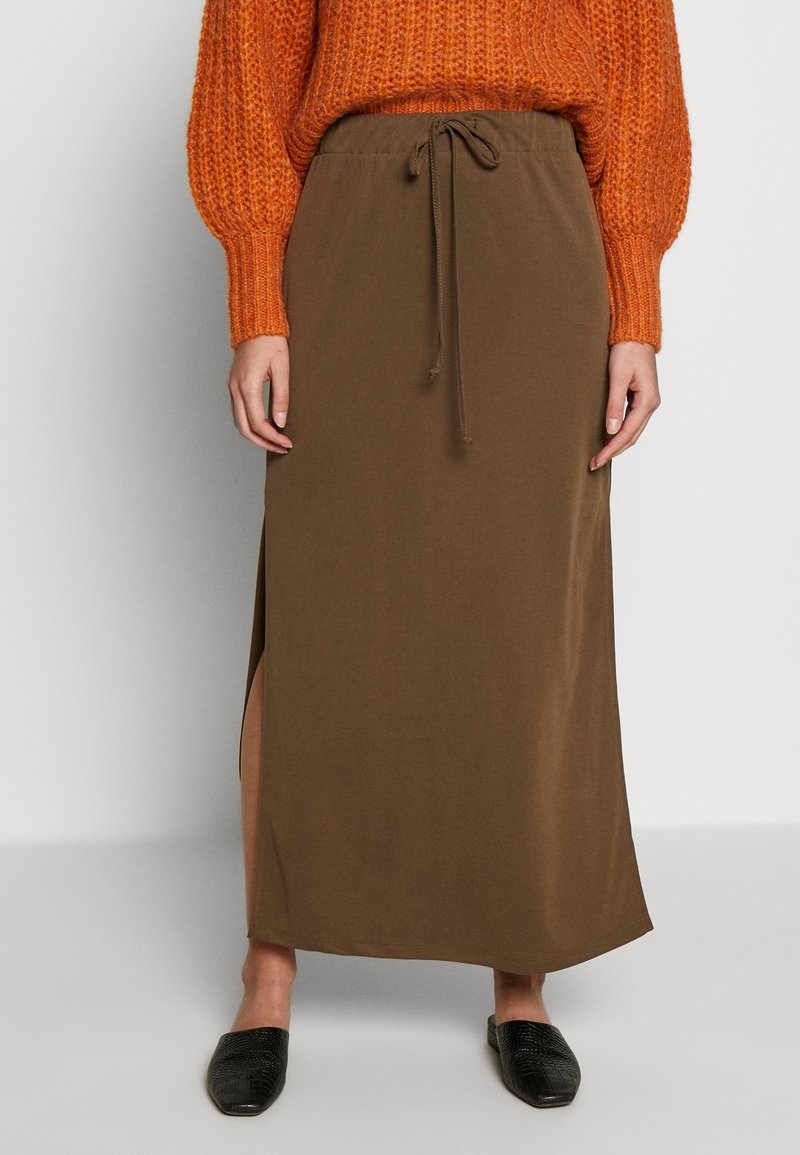 Jupe marron arrivant aux genoux, en tissu lisse, avec une taille à cordon et des fentes latérales. Associée à un pull orange en tricot.