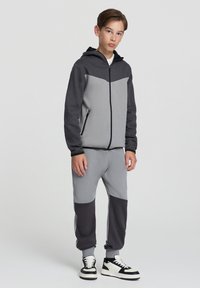 Szary i czarny hoodie z zamkiem, z kontrastowym wzorem i bocznymi kieszeniami, w połączeniu z dopasowanymi joggerami i białymi akcentami w postaci sneakersów.