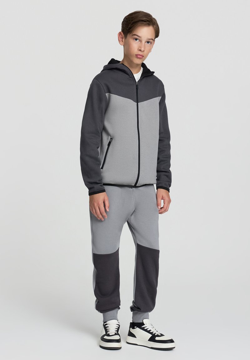 Szary i czarny hoodie z zamkiem, z kontrastowym wzorem i bocznymi kieszeniami, w połączeniu z dopasowanymi joggerami i białymi akcentami w postaci sneakersów.