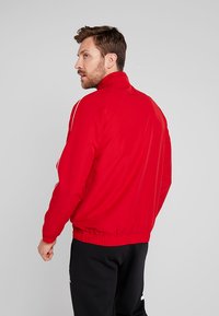 Rote, mit Reißverschluss versehene Sportjacke mit hohem Kragen, elastischen Bündchen und Saum. Verfügt über weiße Streifen entlang der Schultern. Glatte Textur.