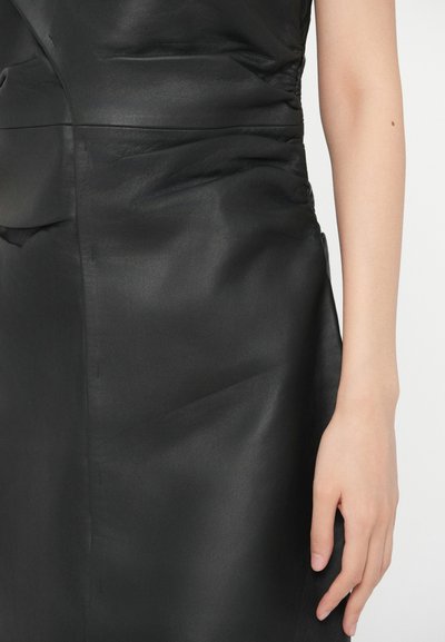 Fekete bőrruha sima textúrával, a szélén redőzött részletekkel és alakhoz illeszkedő siluettel, részben kéz által bemutatva.
