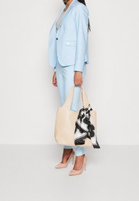 Blazer bleu clair et pantalon assorti avec un sac à main crème orné d'un foulard noir et blanc à motifs. Des talons aiguilles beige complètent la tenue.
