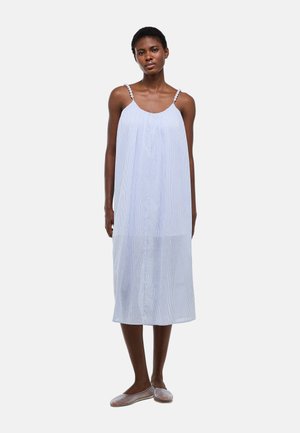 Femme debout portant une robe midi bleu clair sans manches avec de fines bretelles et des chaussures tissées plates sur un fond blanc uni.