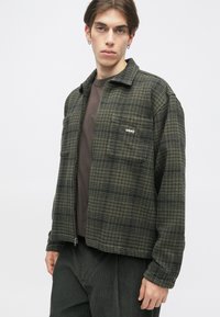 Zeleni karirani flis jakna z zadrgo spredaj, s koničastim ovratnikom in izvezenim logotipom na prsih. Oblačena je čez rjavo srajco in temne hlače.