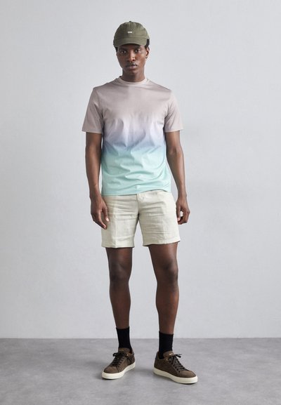 Uomo che indossa una camicia a maniche corte sfumata, pantaloni beige, cappellino verde, calze nere e sneakers marroni, in piedi contro un muro grigio uniforme.