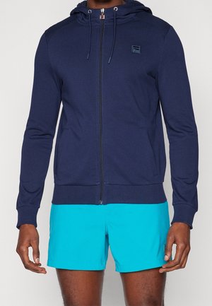 Man draagt een marineblauwe zip-up hoodie met capuchon en turquoise korte broek, staand tegen een effen lichte achtergrond.