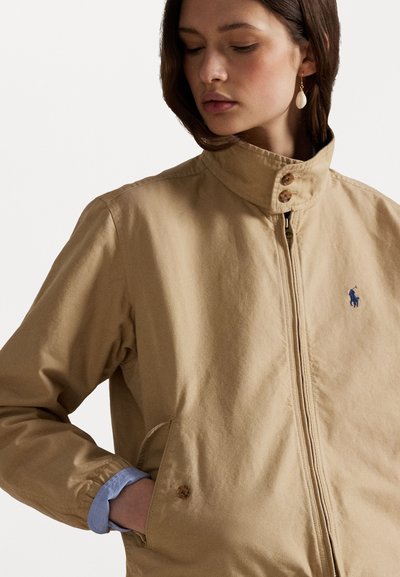 Hellbraune Jacke mit hohem Kragen, Frontreißverschluss und zwei seitlichen Taschen. Verfügt über Holzknöpfe und ein dezentes blaues Logo auf der Brust.