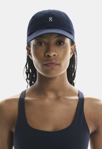 Navy blue cap made of fabric, featuring a curved brim and embroidered logo on the front. The model wears a dark sports tank top.