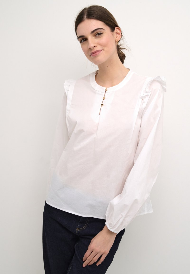 Blusa di cotone bianca con dettagli arricciati sulle spalle, scollo rotondo e tre bottoni decorativi frontali. Maniche lunghe a sbuffo con polsini arricciati.