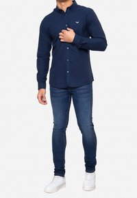 Camisa azul marino de botones con mangas largas y cuello, hecha de tela suave, con un pequeño emblema en el pecho. Se lleva con jeans ajustados y zapatillas blancas.