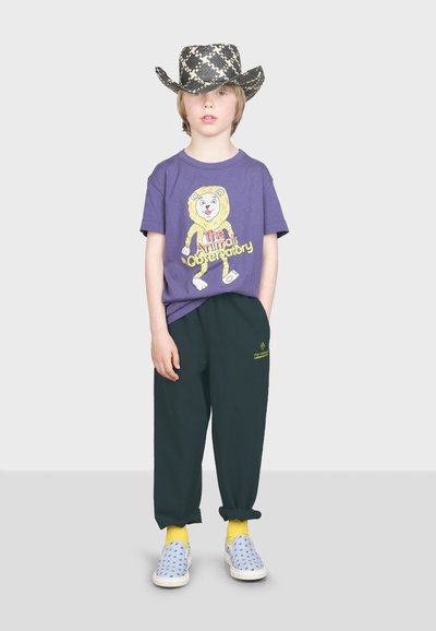 Fialové bavlnené tričko s grafickým zobrazením leva, tmavozelené teplákové nohavice, modré slip-on topánky s výrezmi a čierny klobúk so vzorom kovboja.