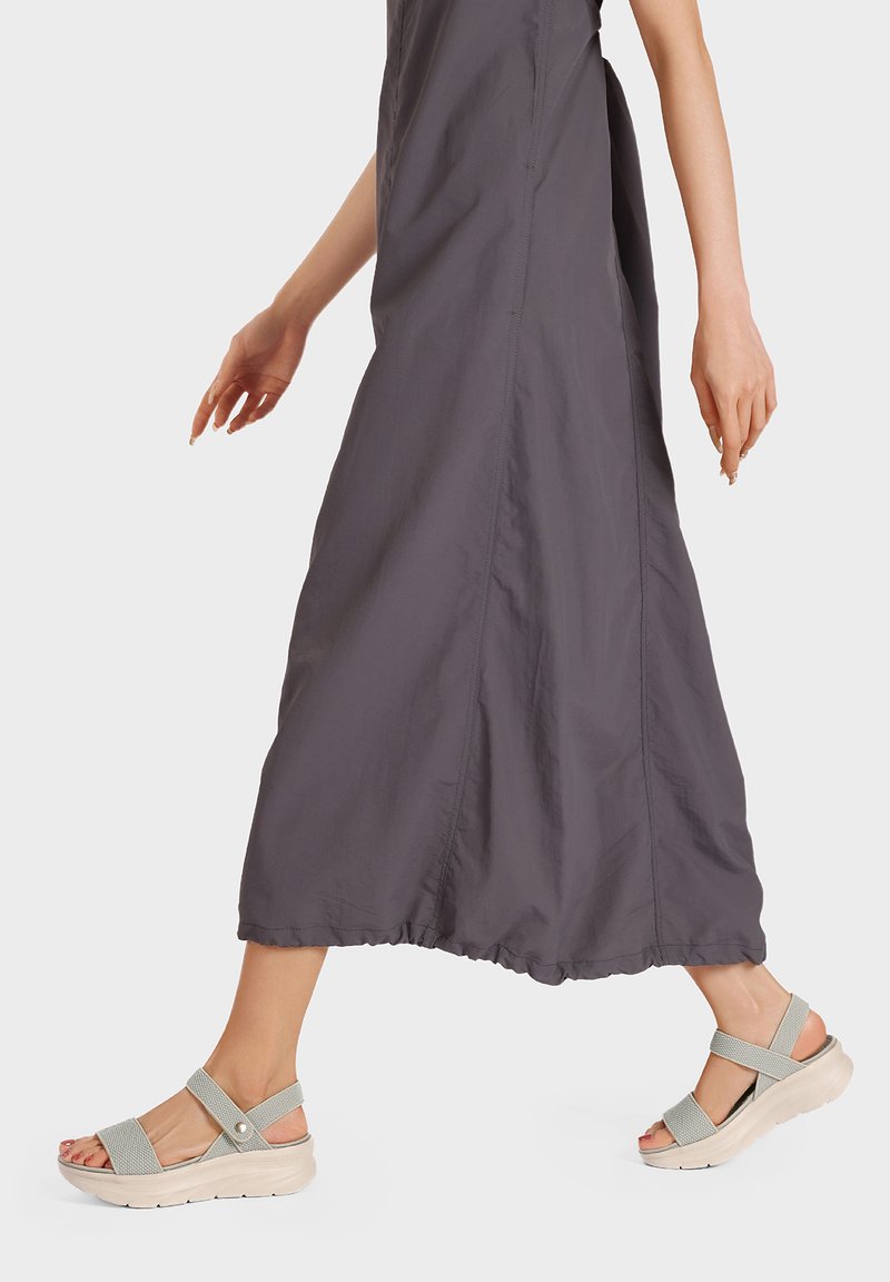 Sandali grigi chiari con cinturini incrociati, punta aperta e suola piatta imbottita. Indossati con un lungo vestito grigio in una posa casual mentre si cammina.