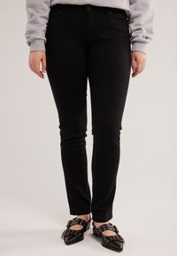 Személy fekete slim-fit nadrágot, szürke hosszú ujjú felsőt és fekete, dupla csattal és fémfüllel díszített hegyes orrú cipőt visel.