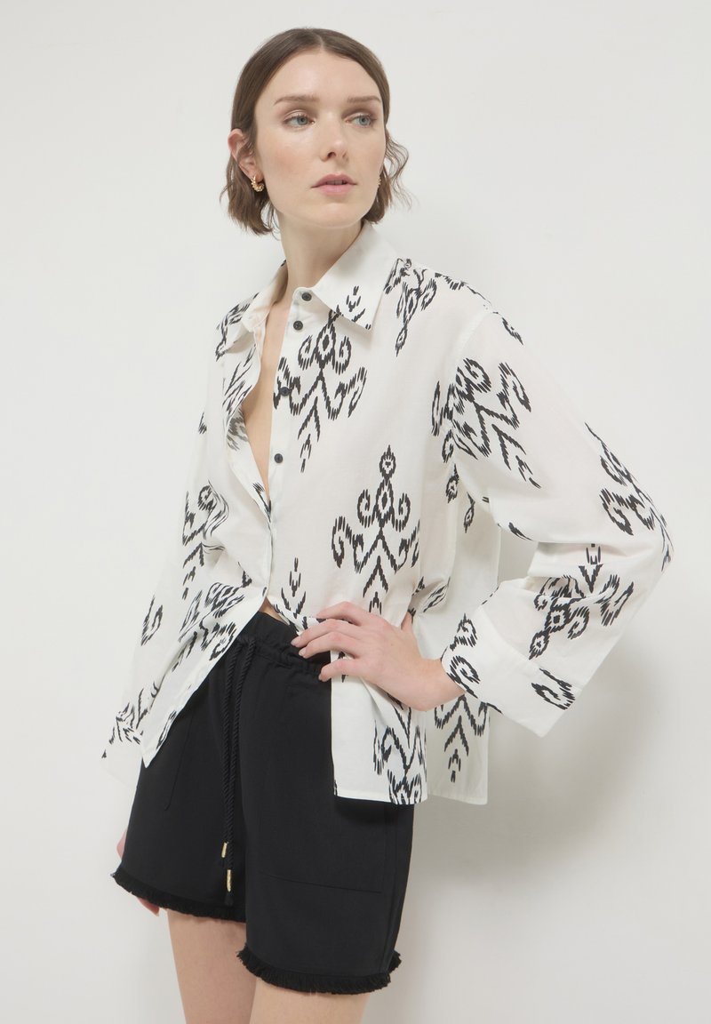 Camicia bianca con pattern ikat neri, maniche lunghe e vestibilità ampia, abbinata a pantaloni corti neri con orlo frangiato e vita elastica.