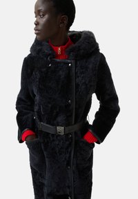 Sort pelsjakke med hætte, der har en belagt talje, trykknapper, sidelommer og røde strikkede manchetter. Blød tekstur og varmt design.