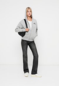 Siva zip-up dukserica s logom, uparena s crnim širom hlačama i bijelim tenisicama. Manekenka nosi malu crnu torbicu preko tijela.