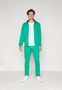 Green tracksuit made of lightweight material, featuring a zip-up jacket and fitted pants. White t-shirt underneath and white sneakers.