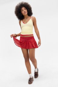 Młoda kobieta z naturalną fryzurą afro, ubrana w żółty top na ramiączkach i czerwoną mini spódniczkę w kropki, trzymająca brzeg spódniczki i patrząca w bok.