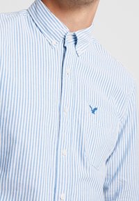 Chemise rayée bleu clair et blanche, boutonnée, avec un col et une poche avant, ornée d'un petit logo brodé bleu sur la poche.