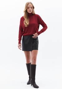 Blusa rossa a maniche lunghe con collo a dolcevita abbinata a una gonna a portafoglio nera in ecopelle e stivali neri con tacco al ginocchio, caratterizzati da una texture liscia.