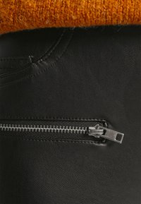 Tecido de couro preto com um acabamento texturizado, apresentando um fecho metálico em prata. Um detalhe em malha laranja parcialmente visível na parte superior.