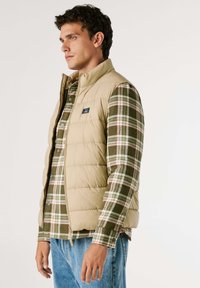 Gilet imbottito kaki con colletto alto, indossato sopra una camicia a quadri verde, rossa e bianca. Il gilet presenta una chiusura con zip e un marchio discreto.