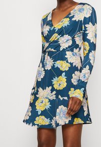Robe portefeuille à motifs floraux en tissu bleu marine avec des manches longues, présentant des fleurs jaunes et violet clair, avec une taille cintrée et une longueur au-dessus du genou.
