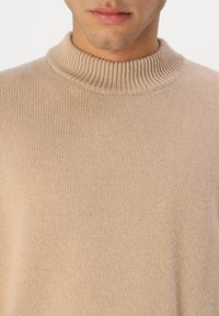 Beżowy sweter z dzianiny z wysokim kołnierzem, teksturowanym materiałem i luźnym krojem, podkreślający miękką, gładką powierzchnię.