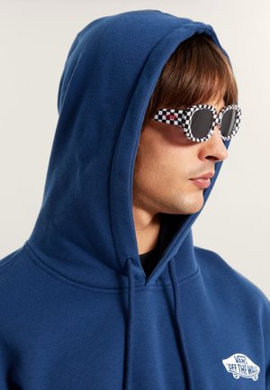 Blauwe katoenen hoodie met een trekkoord in de capuchon, voorzien van een wit logo op de borst. Draagt een bril met een ruitvormig montuur en donkere lenzen.