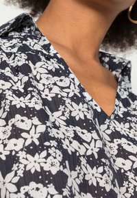 Navy Blumenbluse mit weißen Blumenmustern, weicher Textur, kurzem Kragen und V-Ausschnitt. Auffälliger Nadelstreifen-Hintergrund.