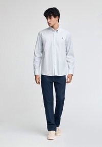 Ljusblå randig skjorta med knappar och långa ärmar, rak krage och broderade logotyper, tillsammans med mörka denimjeans och beige sneakers.