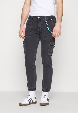 Man draagt zwarte cargobroek met elastische enkels, een blauw sleutelhangerlusje, witte sneakers met zwarte strepen en een wit T-shirt.