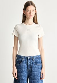 Μπεζ ριμπ κοντομάνικο T-shirt που διαθέτει ένα μικρό λογότυπο στο στήθος, σε συνδυασμό με μπλε τζιν παντελόνι που παρουσιάζει ένα επαναλαμβανόμενο σχέδιο κειμένου.
