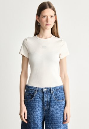 Beige ribbade t-paita lyhyillä hihoilla, jossa on pieni logo rinnassa, yhdistettynä sinisiin farkkushortsiiin, joissa on toistuva tekstikuosi.
