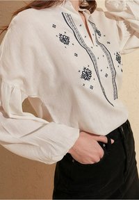 Hvid bluse med sort blomstermønster på brystet, lange pufærmer og en hånd stoppet i lommen på et par sorte højhalsede bukser.