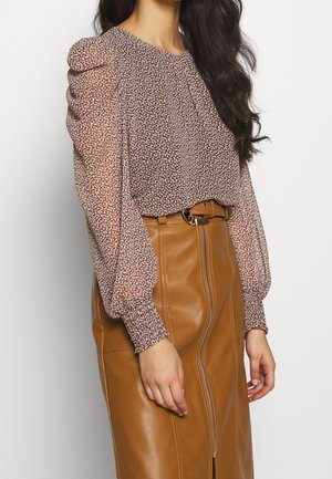 Femme portant un chemisier à manches longues fleuri avec des épaules bouffantes, rentré dans une jupe en cuir marron taille haute avec une fermeture éclair devant et une ceinture.