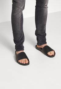 Sandalias negras tipo slide con una amplia correa plana y punta abierta, combinadas con jeans de mezclilla gris. Las sandalias tienen una textura suave y un diseño elegante.