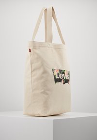 Beige canvas shoppingväska med två handtag, prydd med en svart Levi's-logotyp omgiven av blommönster på framsidan. Slät textur.
